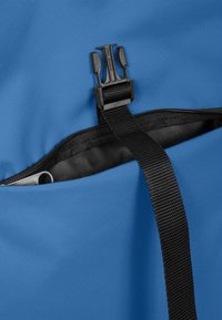 Blå tygväska med en texturerad yta, försedd med en svart dragkedja och ett bärrem. Innehåller en plastspänne för säker stängning.