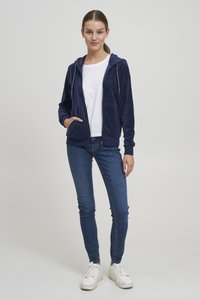 Tmavomodrá mikina na zips z mäkkej látky s dvoma prednými vreckami, kombinovaná s priliehavými modrými džínami a bielymi teniskami. Jednoduchý dizajn s kapucňou.