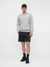 Homme portant un sweat-shirt gris à manches longues, un short noir, des chaussettes noires, des baskets noires et une casquette de baseball noire, debout devant un fond blanc uni.