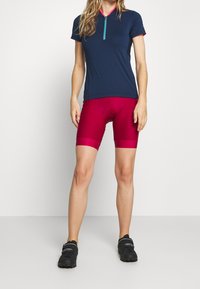 Jersey de ciclismo de manga corta azul marino con cremallera y cuello rosa, combinado con pantalones cortos de ciclismo ajustados de color magenta. Zapatos de ciclismo negros completan el conjunto.