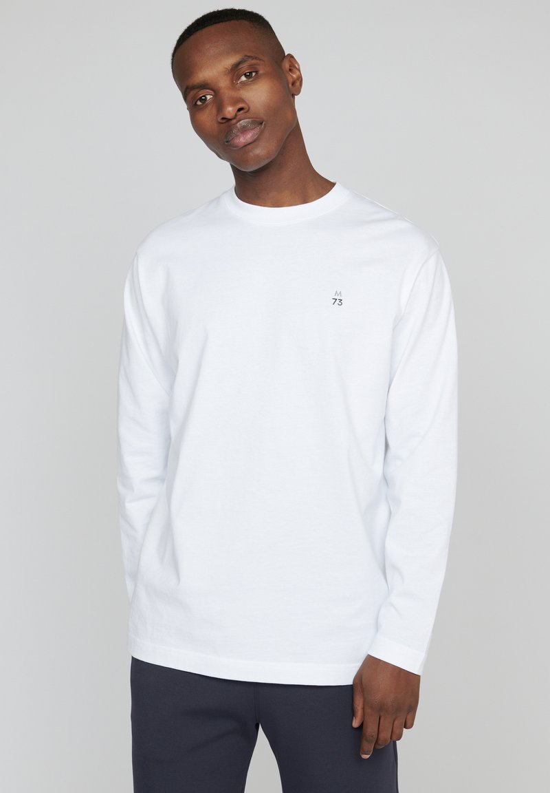 Balta ilgomis rankovėmis marškinėliai iš medvilnės, su mažuoju pilku logotipu kairėje krūtinės pusėje. Paprastas, apvalios kaklo iškirpės modelis su įprastu sijonu.