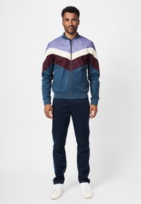 Track jacket featuring a zip closure, color-blocked design in purple, white, burgundy, and teal. Paired with navy pants and white sneakers.