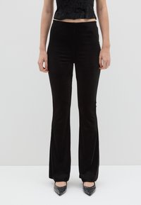 Pantaloni a zampa d'elefante in velluto nero con una texture morbida, dotati di vita alta e design aderente, abbinati a tacchi neri con borchie.