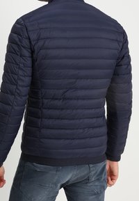 Marineblå pufjakke med horisontale sømme og ståkrave. Har elastiske manchetter og bund. Letvægtsmateriale, slankt design.