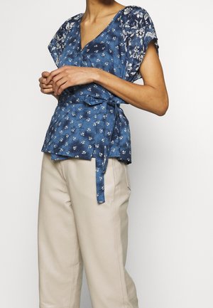 Frau trägt eine blaue Wickelbluse mit floralem Muster und kurzen Ärmeln sowie beige hochtaillierte Hosen, die Hände locker vor dem Körper verschränkt.