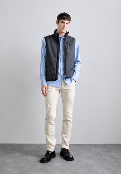 Black puffer vest over a light blue button-up shirt, paired with cream-colored pants and black shoes, standing against a neutral background.