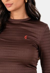Vrouw in een bruine top met lange mouwen van textuur, voorzien van een klein rood geborduurd ontwerp, gouden hoopoorbellen en loshangend donker haar.