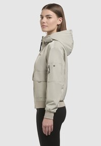 Light beige hooded jacket with a smooth texture, zippered pockets, and ribbed cuffs. Slim fit with a simple design and visible logo accent.
