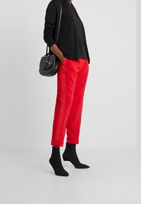 Rote, taillierte Hose mit Seitenstreifen, kombiniert mit einem schwarzen Pullover und hohen Sockenstiefeln. Die Accessoires umfassen eine kleine schwarze Handtasche.