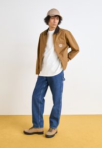 Chaqueta de lona marrón con un collar marrón, camiseta blanca, vaqueros azules y botas de senderismo beige. El entorno presenta un suelo amarillo.