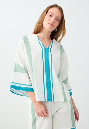 Jeune femme portant un haut blanc ample et un pantalon avec des rayures verticales turquoise et vertes, posant devant un fond clair uni.