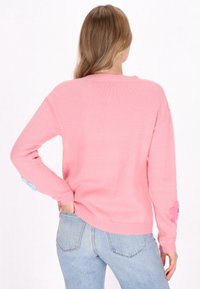 Suéter de punto rosa con mangas largas, que presenta un escote acanalado y coloridos apliques florales en los puños. Combinado con jeans de color azul claro.