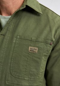 Green canvas shirt with a prominent chest pocket and branded patch. Features a classic collar and textured fabric.