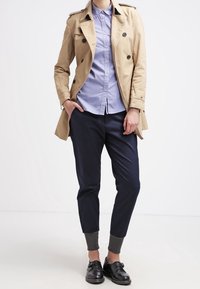 Cappotto beige sopra una camicia azzurra con bottoni, pantaloni blu navy con risvolti grigi e scarpe nere. Il cappotto presenta bottoni doppi.