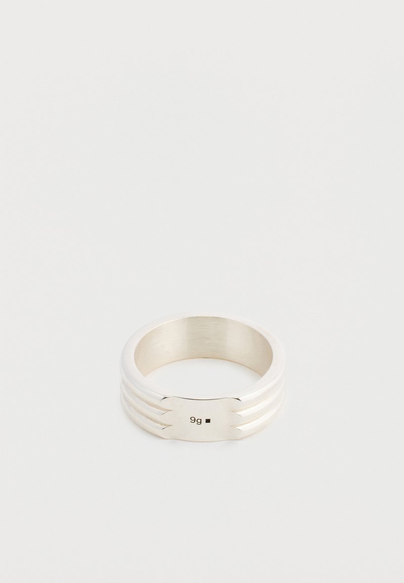 Une bague ton argent avec une large plaque frontale marquée « 9g » et deux rainures de chaque côté, présentée sur un fond clair uni.