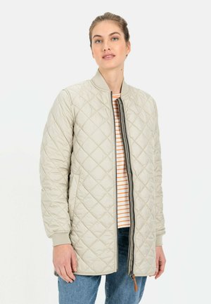 Helle beige, gesteppte Jacke mit Reißverschluss, gerippten Bündchen und einem runden Kragen. Innen ist ein gestreiftes Hemd zu sehen, das mit blauen Jeans getragen wird.