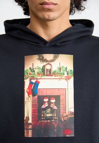 Sudadera negra con un gráfico de una escena navideña que incluye una chimenea, calcetines colgados y zapatillas rojas y blancas con gorros de Santa.