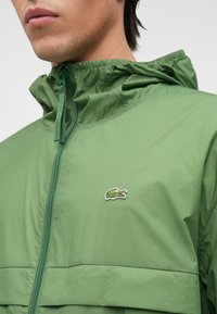 Πράσινο ελαφρύ μπουφάν με κλείσιμο φερμουάρ, κουκούλα και κεντητό λογότυπο κροκόδειλου. Διαθέτει υφή υφάσματος και ελαστικές μανσέτες.