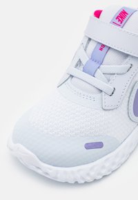 Zapatilla deportiva de color gris claro con parte superior de malla, cuello acolchado y acentos en rosa. Tiene una suela texturizada y un detalle de logo en púrpura.
