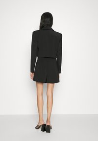 Kvinna klädd i en svart kort blazern och högmidjade shorts, stående vänd mot en vit vägg, med svarta klackmules på fötterna.