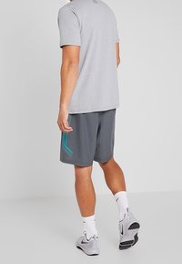 Muž oblečený v světlém šedém tričku, tmavě šedých sportovních šortkách s tyrkysovým designem, bílých kotníkových ponožkách a šedých teniskách Nike, stojící proti bílému pozadí.