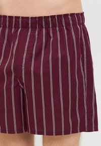 Costume de baie burgundy cu dungi verticale albe, material ușor, talie elastică și un singur buzunar frontal.