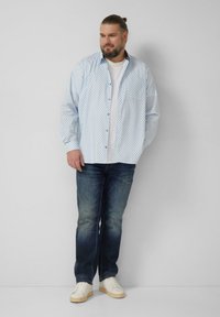 Weißes Hemd mit blauen geometrischen Mustern, kombiniert mit blauer Jeans und weißen Turnschuhen. Mit langen Ärmeln und lockerem Schnitt.