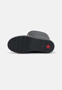 Botte en caoutchouc noire avec une semelle texturée, un bout arrondi et un dessus lisse. Présente un accent rouge près du talon et un motif de semelle robuste.