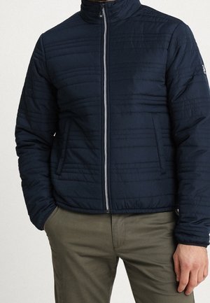 Mężczyzna ubrany w granatową pikowaną kurtkę na zamek błyskawiczny z bocznymi kieszeniami oraz oliwkowe spodnie, stojący na jednolitym tle.