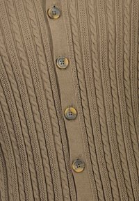 Beige-neulepusero, jossa on pystysuorat palmikkokuvioinnit ja neljä pyöreää, marmorikuvioista nappia etumuksessa.