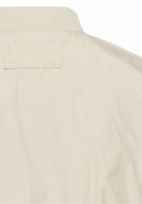 Dos d'une veste beige montrant un col texturé et une étiquette en tissu cousue indiquant "Camel Active" près de l'encolure.