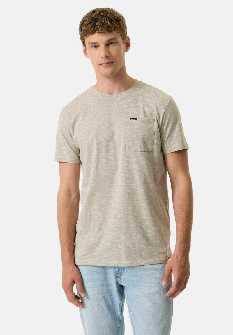 Joven con cabello corto y rizado, vistiendo una camiseta beige de manga corta con bolsillo y jeans azul claro, de pie contra un fondo liso.