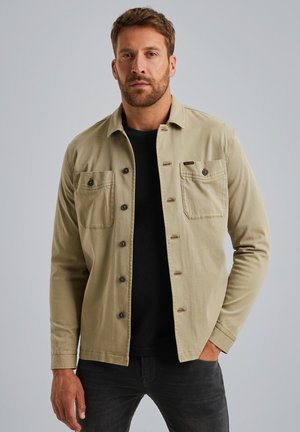 Camisa de botones de algodón beige con dos bolsillos en el pecho, botones oscuros y un collar, llevada sobre una camiseta negra, combinada con jeans oscuros.
