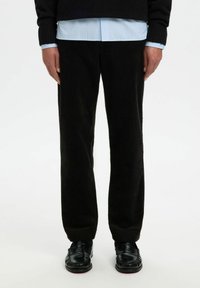 Isik, kellel on seljas mustad velvetpüksid, mustad nahkloafersid ja kihiline riietus, mille alla on pandud helesinine särk musta sviitri all.