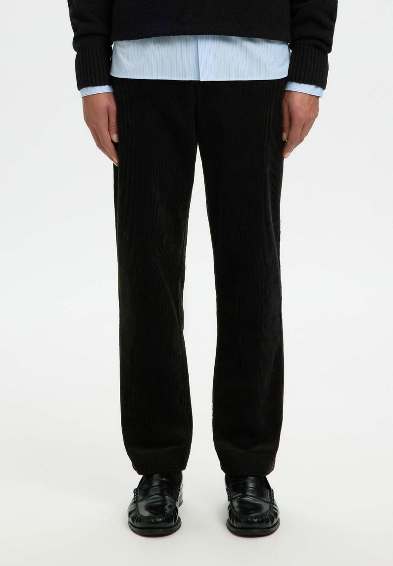 Isik, kellel on seljas mustad velvetpüksid, mustad nahkloafersid ja kihiline riietus, mille alla on pandud helesinine särk musta sviitri all.