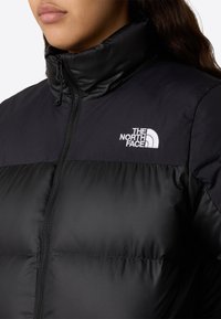 Svart dunjakke med høy krage, glidelås, quiltet tekstur og hvit "The North Face"-logo på øvre venstre side.