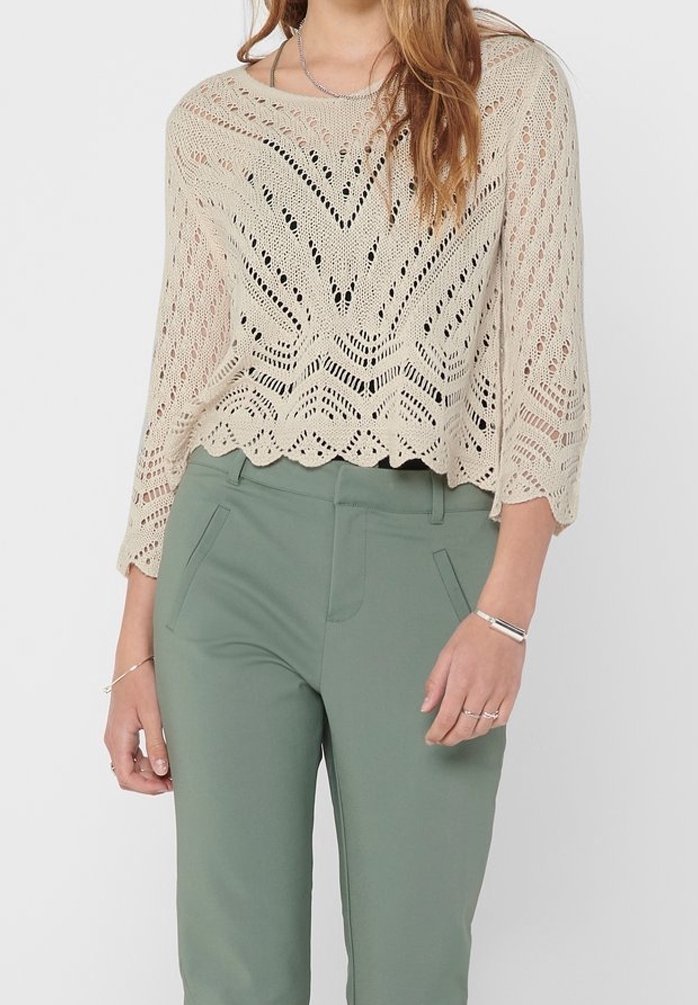 Femme portant un haut beige en crochet à manches longues et un pantalon vert taille haute, montrant des bijoux à ses mains et poignets.