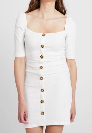 Femme portant une robe blanche moulante côtelée avec des manches jusqu’au coude et de gros boutons marron au centre devant.