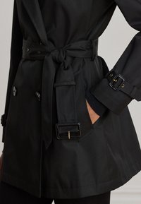 Zwarte trenchcoat met een gestrikte tailleriem, gespen op de manchetten, dubbelrijige knopen en een hand in de zijzak.