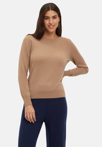 Ronde hals, lange mouwen trui in camel-kleurige gebreide stof; heeft een geribde zoom en manchetten; gedragen met donkerblauwe broek.