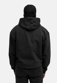 Schwarzer übergroßer Hoodie aus strukturiertem Stoff, ausgestattet mit einer großen Kapuze und gerippten Bündchen, getragen von einer Person, die mit dem Rücken zum Betrachter steht.