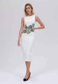 Donna in un vestito midi senza maniche bianco con ricami floreali neri in vita, che indossa tacchi alti neri e gioielli minimi.