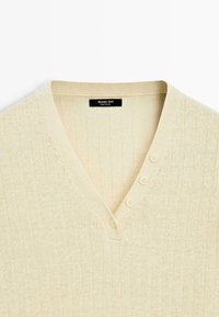 Cardigan in maglia a costine color crema con scollo a V, quattro piccoli bottoni lungo la scollatura e un'etichetta "Massimo Dutti" all'interno del colletto.