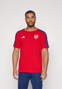 T-shirt rouge à manches courtes avec col rond, présentant un logo blanc d'Arsenal sur la poitrine et des accents bleus le long des épaules.