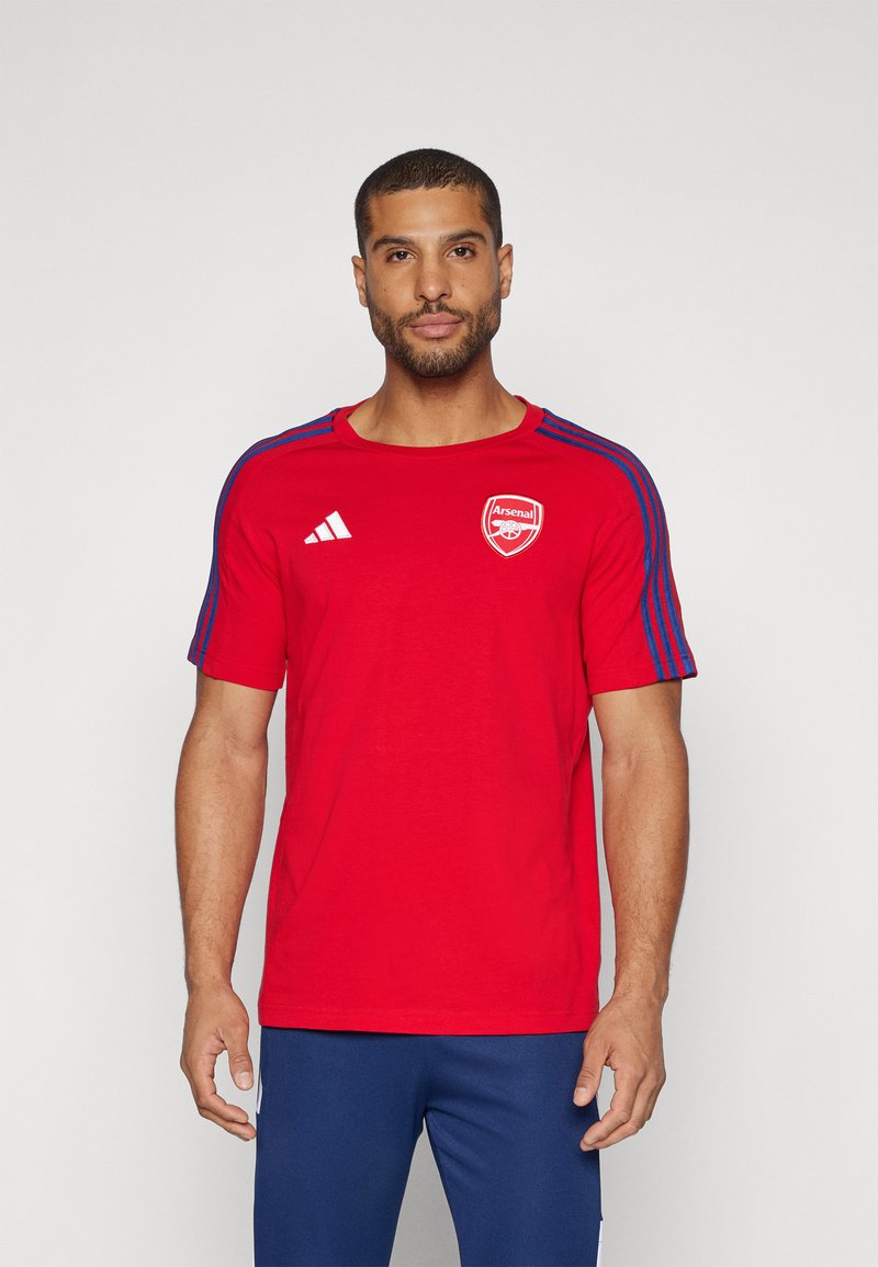 T-shirt rouge à manches courtes avec col rond, présentant un logo blanc d'Arsenal sur la poitrine et des accents bleus le long des épaules.
