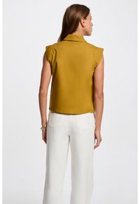 Mosterdgele mouwloze blouse met kraag, cropped ontwerp en gladde stof; gecombineerd met witte high-waisted broek met achterzakken.