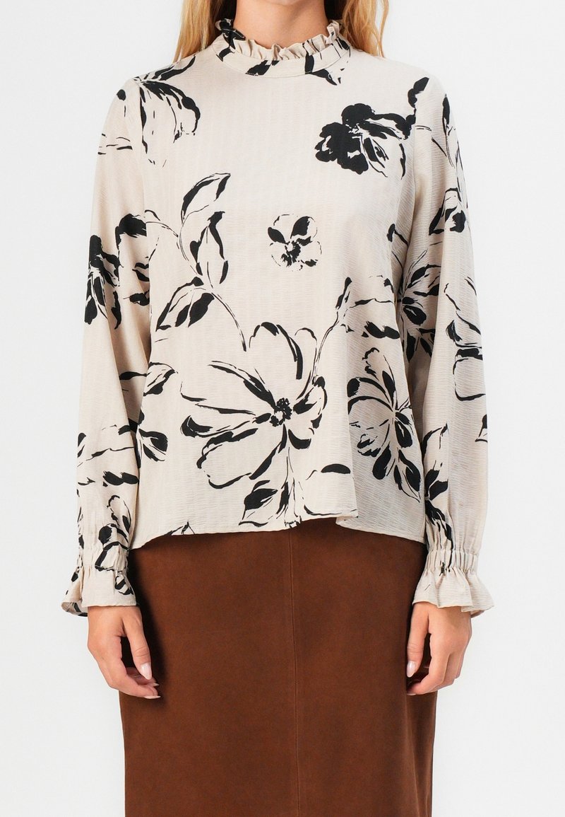 Langærmet bluse i beige stof med sort blomsterprint, flæsekrave og rynkede manchetter. Sammen med en brun nederdel.