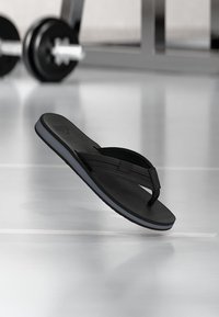 Μαύρο σανδάλι τύπου φλιπ-φλοπ που αιωρείται πάνω από το αντανακλαστικό δάπεδο γυμναστηρίου, με θολό βάρος ανύψωσης στο φόντο.