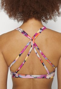Henkilön selkä, joka käyttää moniväristä urheil brasia, jossa on ohuet ristikkäiset hihnat ja kiharat hiukset.