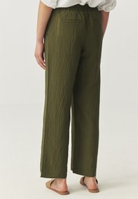 Pantalons larges en vert olive, en tissu texturé, avec une taille élastique et deux poches arrière, associés à des sandales marron.